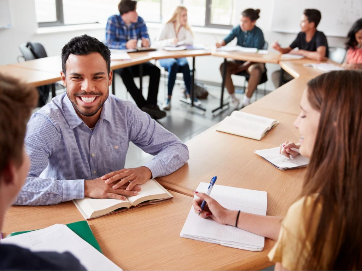 male teacher sitting with students at desk smiling in discussion